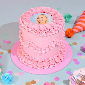 Vintage Photo Cake by Crumbs & Doilies with pink icing and edible printed photo on top, set among party decorations.